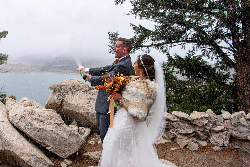 Man pops a bottle of champagne while his bride cheers next to him after their intimate wedding ceremony in Colorado. 