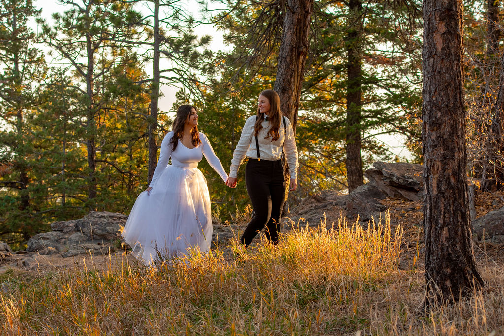 Two women walk hand in hand through a sunlit forest. One wears a flowing white dress, the other dark pants and suspenders. Both are unique and practical elopement outfits for the mountain wedding. The scene is warm and serene.