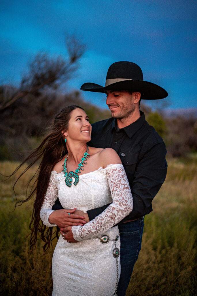 A couple embraces in a grassy field at dusk. The woman, in a white lace dress and turquoise jewelry to spice up her elopement outfit, she smiles up at the man in a black shirt and cowboy hat. Romantic and serene ambiance.