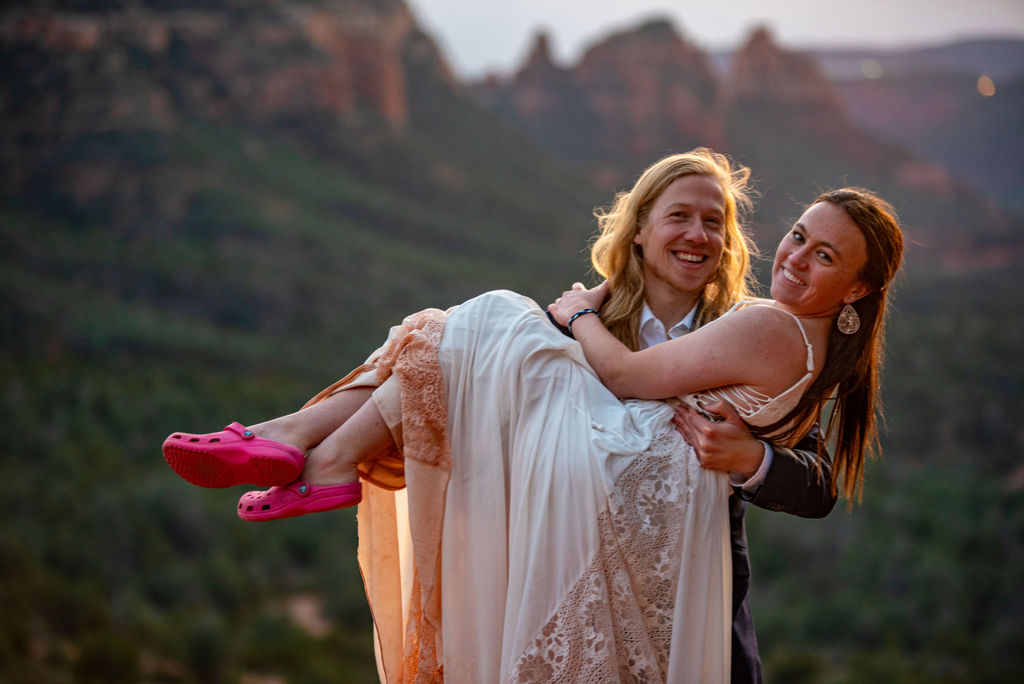 A joyful couple is posed outdoors, with the man carrying the woman who wears a white dress and pink Crocs. A very unique elopement outfit. A scenic, blurred mountain backdrop adds to the lively mood.
