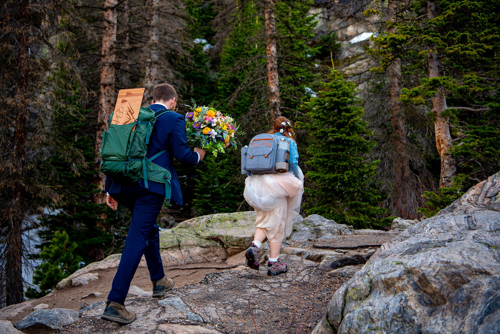A couple hikes uphill in their elopement attire in a forest, wearing backpacks. The man in a suit carries a bouquet, while the woman in a dress wears hiking boots, conveying adventure and joy.