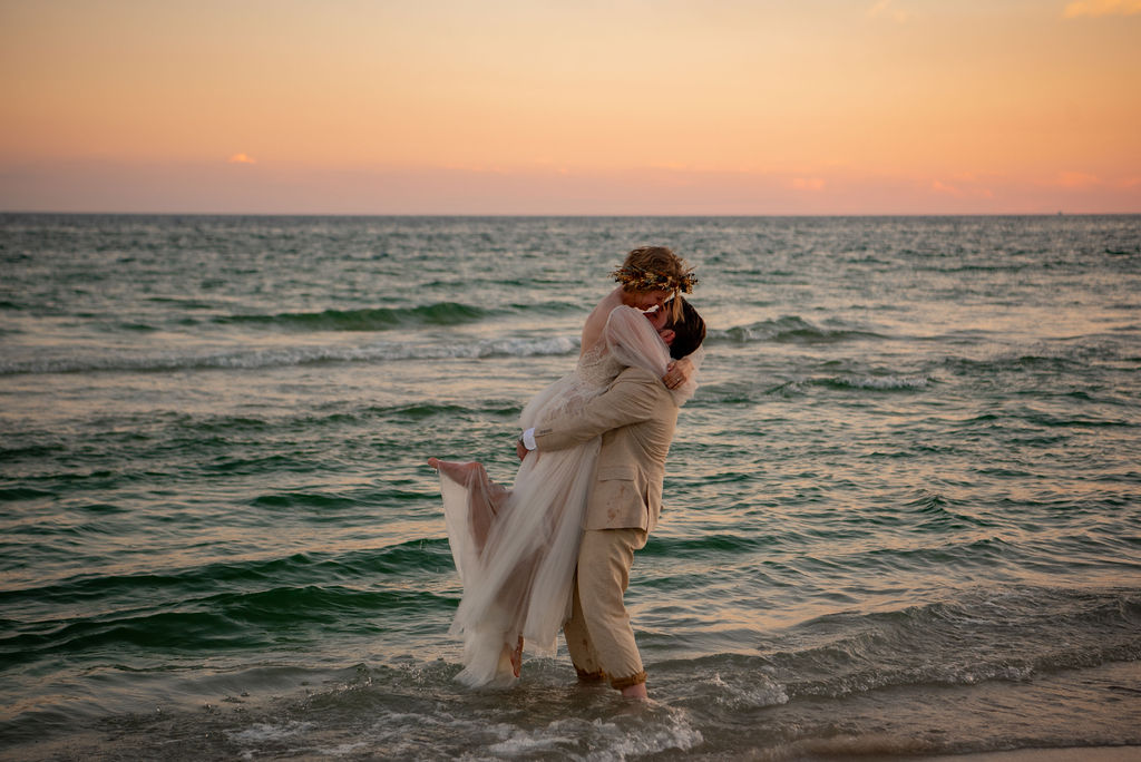 A bride and groom embraces in a joyful moment on a beach as the sun sets, casting a warm, orange glow over the ocean. The bride wears a flowing dress and they are standing in the water. 