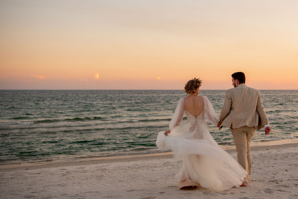 Bride and groom walk hand in hand on a beach at sunset. The brides dress flows behind her. The groom is in a tan suit. Perfect for a beach elopement. The sky glows orange, reflecting on gentle waves. The scene is romantic and serene.
