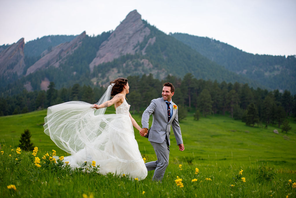 A newlywed couple joyfully runs hand in hand across a vibrant green meadow in Boulder Colorado. The bride's flowing dress contrasts the groom's gray suit, with mountains in the background. It is a perfect balance of their elopement attire and the landscape. 