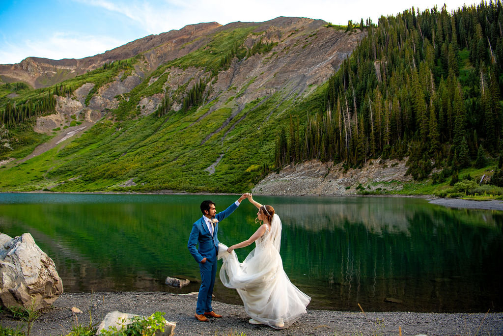 A bride and groom dance in their elopement outfit by a tranquil lake, framed by lush mountains and a clear blue sky. The scene is romantic and serene, with vibrant greenery.