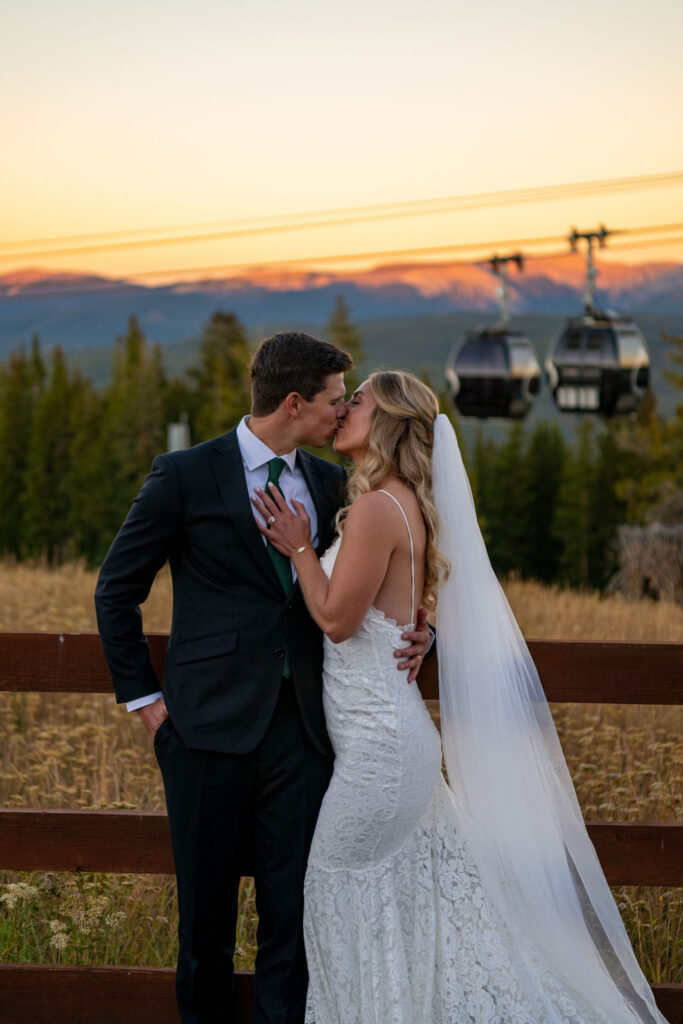 A bride and groom share a kiss in front of a wooden fence during sunset. She wears a white dress and veil; he wears a dark suit. The perfect mountain elopement outfit. Mountains and gondolas are visible in the background.