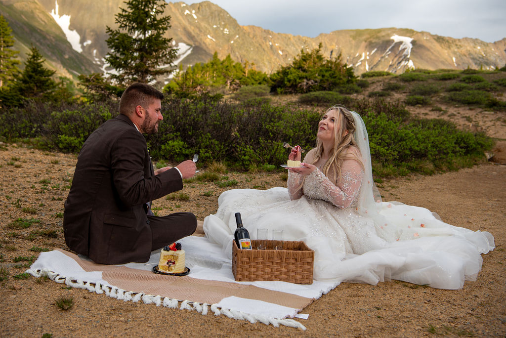 Bride and groom have a picnic on a blanket in a mountainous landscape in their elopement outfits. The bride in a white gown and the groom in a suit share cake, creating a joyful scene.