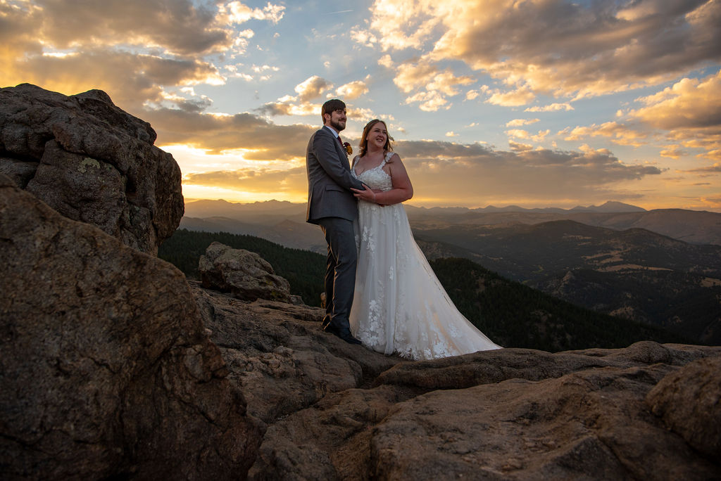 Bride and groom stand in their elopement outfit on a rocky cliff at sunset, with vibrant clouds and distant mountains. They appear joyful and serene, embracing the scenic view.