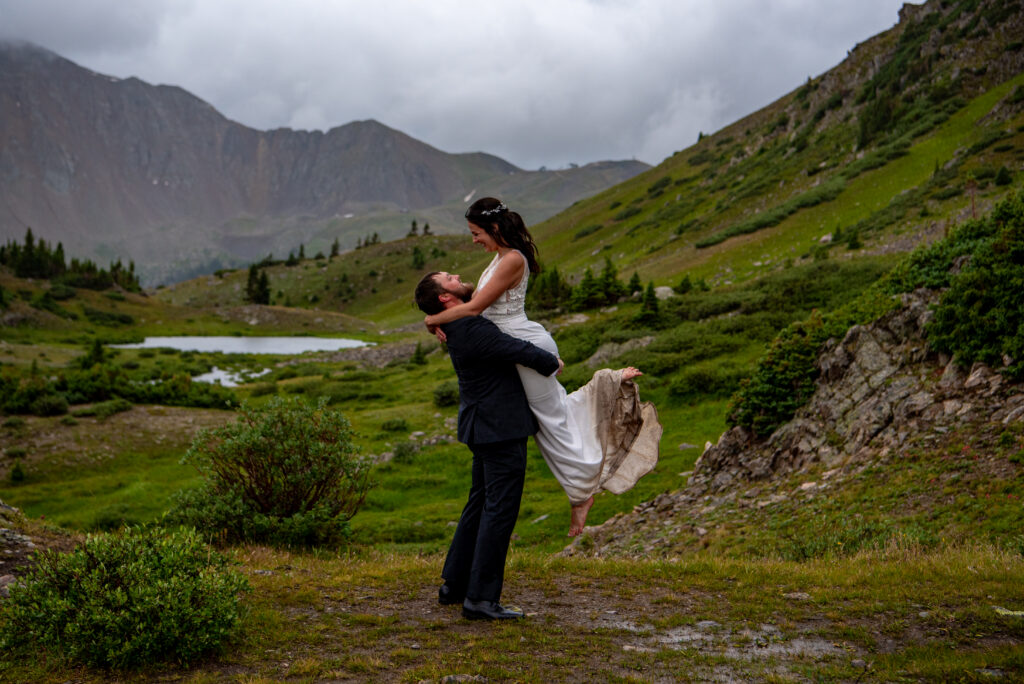 Bride and groom joyfully embrace, groom lifts the bride amid lush, green mountains with cloudy skies and a distant lake, evoking romance and natural beauty. Their elopement outfit is dirty from the mud, but it is elegant and romantic. 