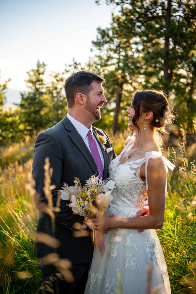 A joyful couple on their wedding day stands in a sunlit meadow. The groom wears a suit and purple tie, and the bride is in a lace dress, holding a bouquet which accents her elopement outfit perfectly