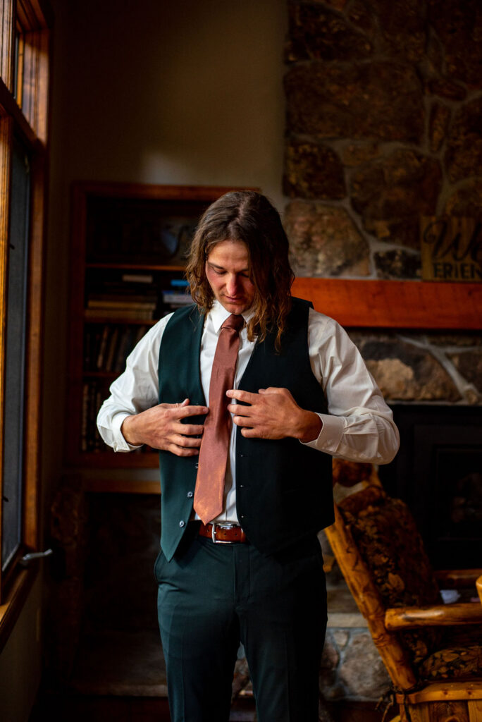 A man in an elopement outfit adjusts his tie in a rustic room with stone walls and a wooden rocking chair. The atmosphere is warm and contemplative.