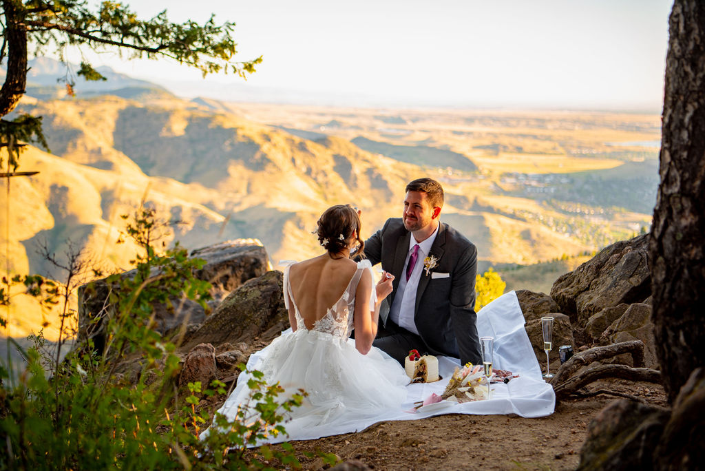 Bride and groom share a picnic on a cliffside, surrounded by scenic mountain views. The bride wears a white dress; the groom a dark suit, a perfect elopement outfit conveying romance.