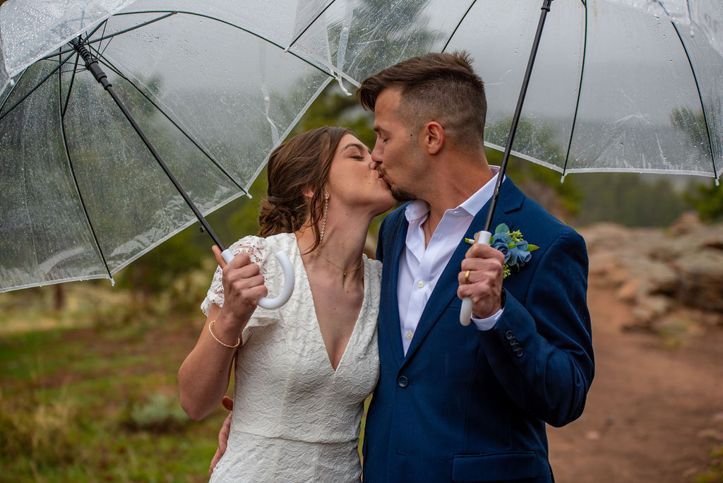 A couple wearing their elopement outfits, kiss under clear umbrellas in a rainy outdoor setting. The mood is romantic and joyful, with a blurred natural background.