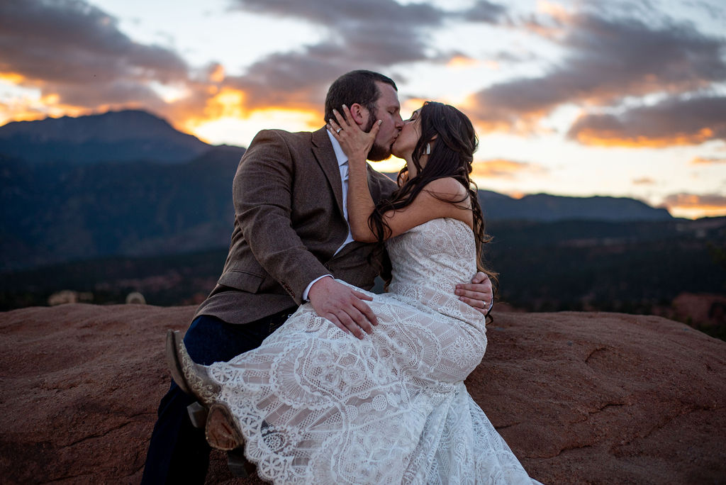 Married couple sit on a rock and share a kiss while the sun sets behind them.