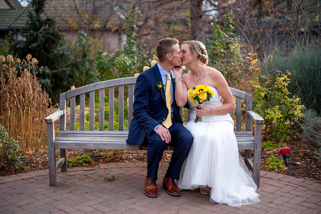 Bride and groom share a kiss while sitting on a bench in the Denver Botanic Gardens