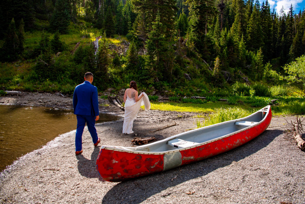 a bride and a groom are exploring the lake shore and looks at a red canoe