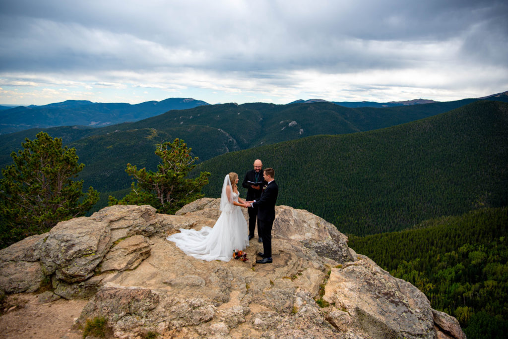 a bride, groom and officiant stand on the edge of a cliff overlooking a Colorado mountain range while the officiant marries the couple. 