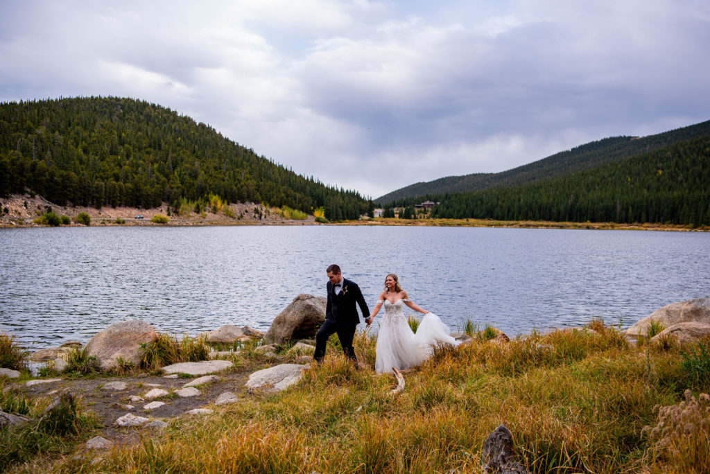 a bride and a groom hold hands and walk through a field with a lake in the background.