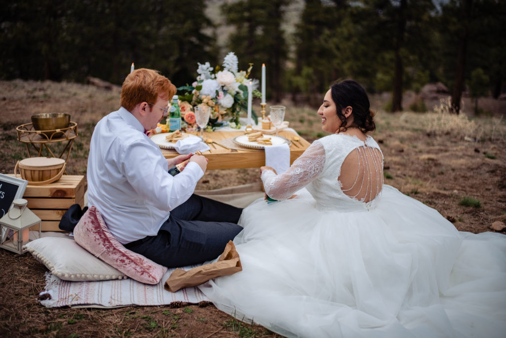 a bride and a groom sit on the ground in their wedding attire and enjoy a luxury picnic with sandwiches and cupcakes