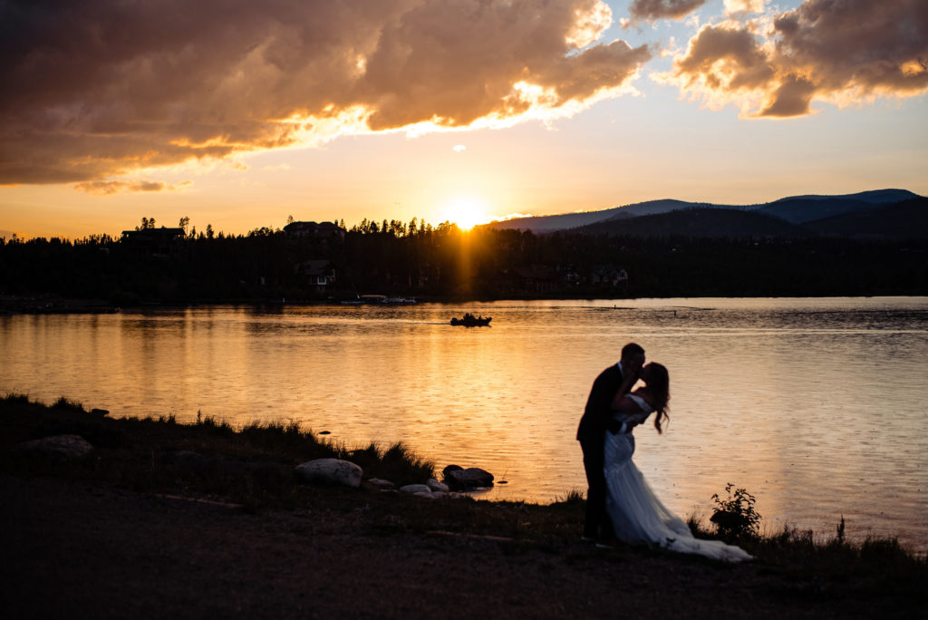 a bride and a groom kiss at sunset by a lake in Grand Lake Colorado while a boat drives by on the lake.