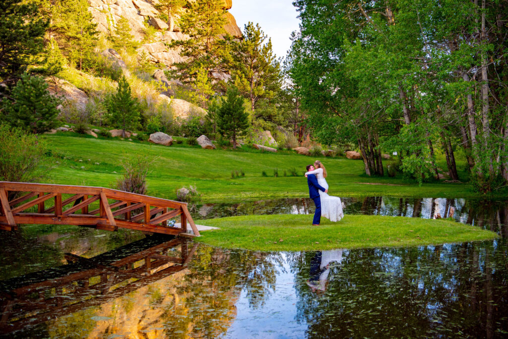 groom holds up his bride in her wedding dress next to a pond with a bridge at Black Canyon Inn for their wedding