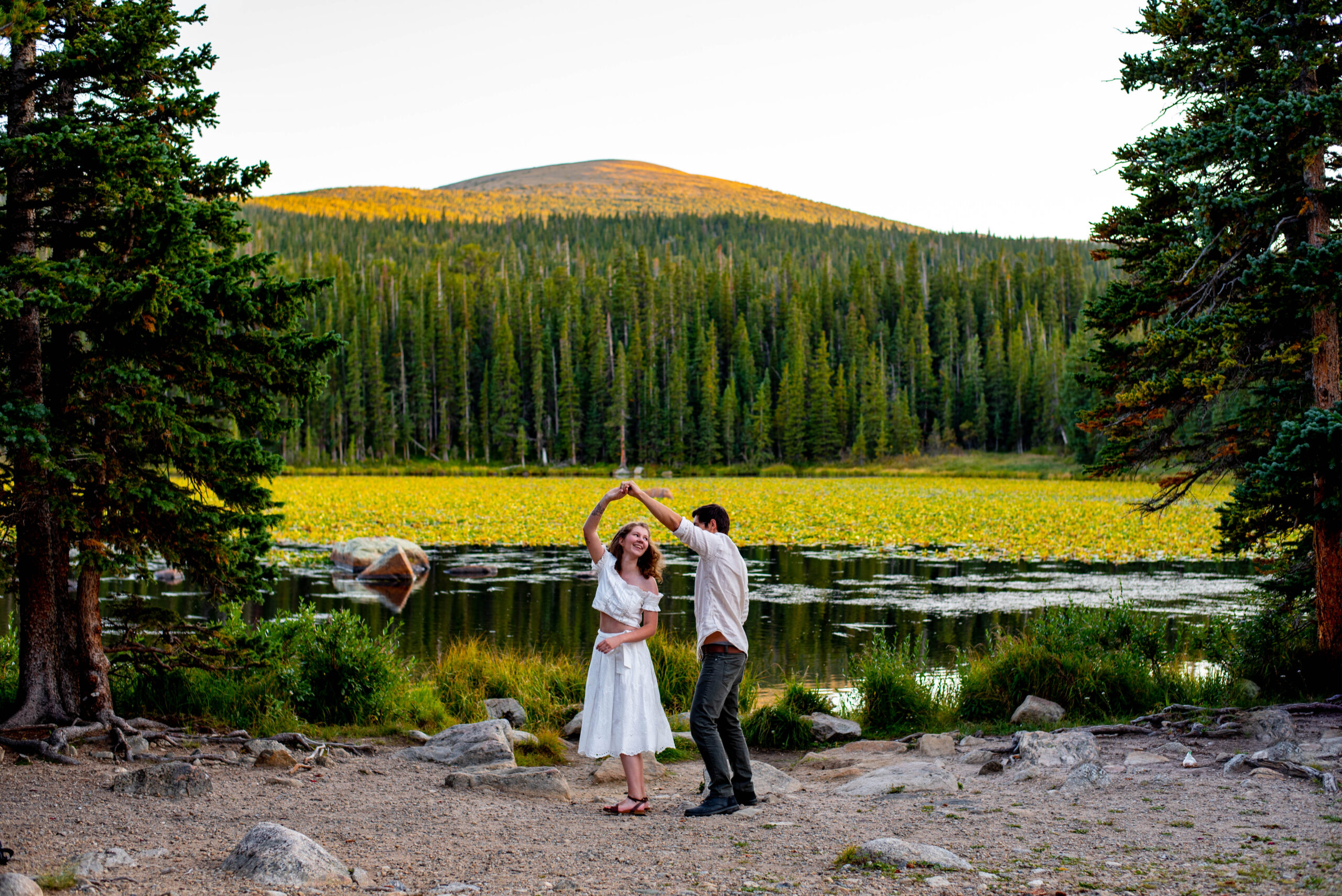 a Colorado engagement photographer captures an image of a man spinning a woman in a white dress in front of an alpine lake.