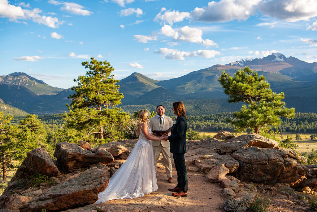 bride and groom say their vows in Rocky Mountain National Park. Bride and groom are holding hands and the officiant stands between them. The sun is shining and there are mountains in the background. 