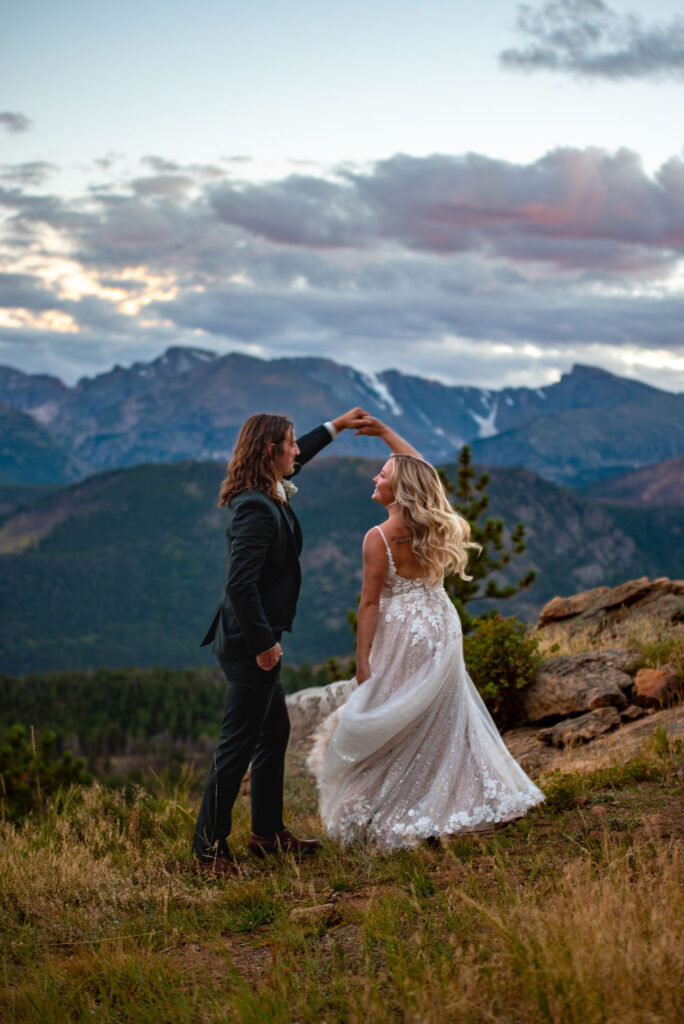 A groom is spinning his bride who is in a white wedding dress on the top of a mountain. The couple is dancing and there are mountains behind them that are turning blue as the sunsets to the right. 