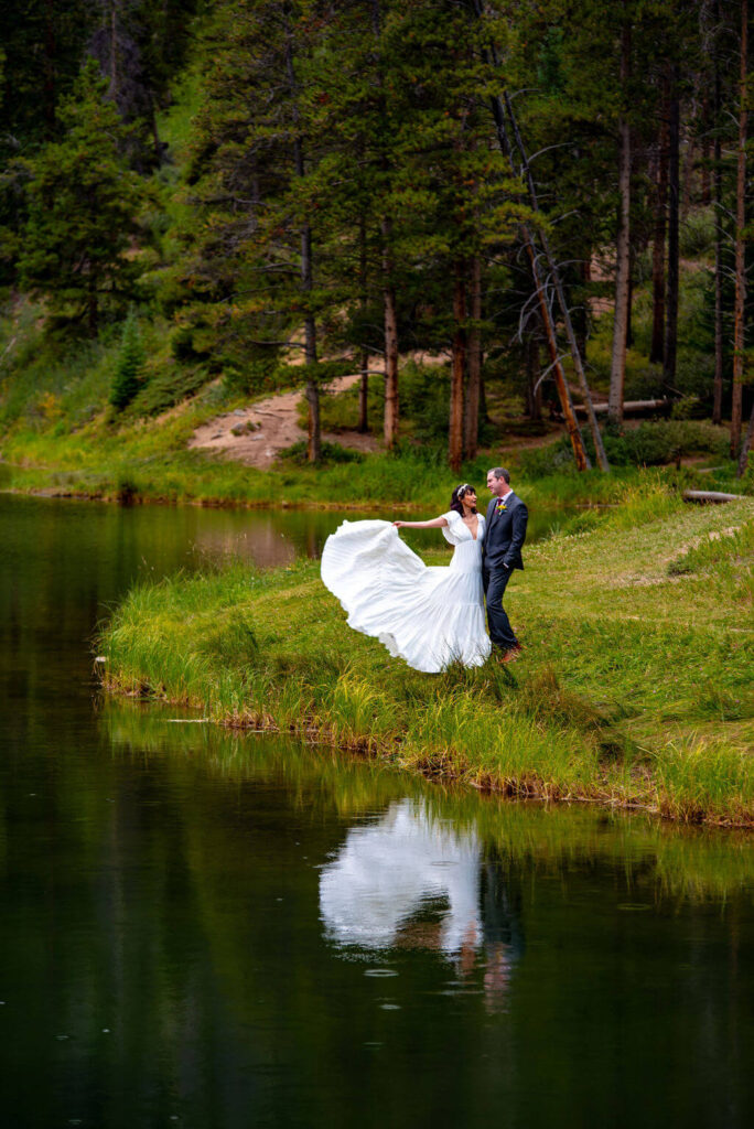A bride and groom are standing on the edge of a lake in a patch of grass. The bride is lifting up the train of her dress and you can see the reflection of the couple in water. Behind the couple is thick green forest.