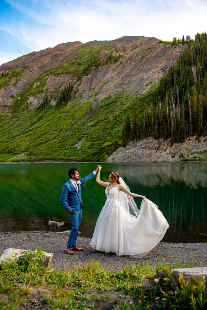 Bride and groom are dancing in front of a lake with mountains behind them. The bride is spinning and throwing out the train of her wedding dress. 
