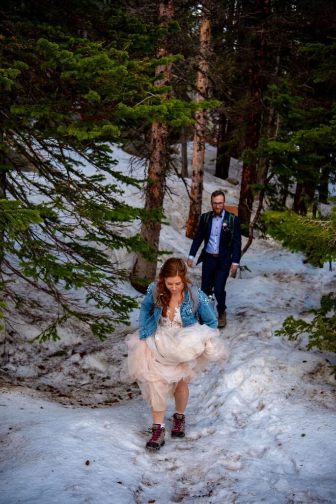A bride in a wedding dress and the groom in a wedding suit are walking through the forest. There is snow on the ground and they are hiking up  a hill. 