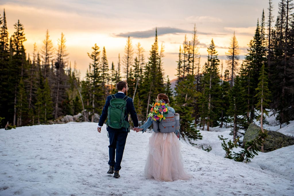 Bride and groom are both wearing backpacks and holding hands as they walk across a snowy hill. They are walking away towards the treeline with a sunset in the background.
