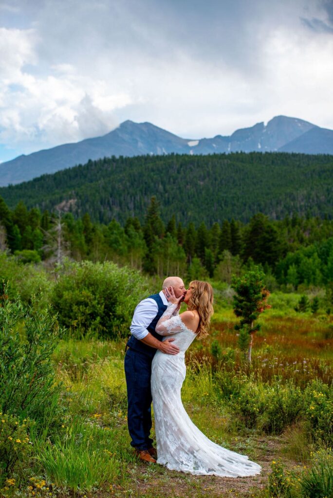 A bride in her wedding dress kisses the groom in a green field surrounded by trees and mountains from Rocky Mountain National Park in the background. 