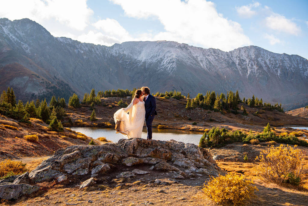 A man and a woman are standing on top of a rock with a snow capped mountain range and a lake in the background. The couple is kissing and they are wearing their elopement attire. 
