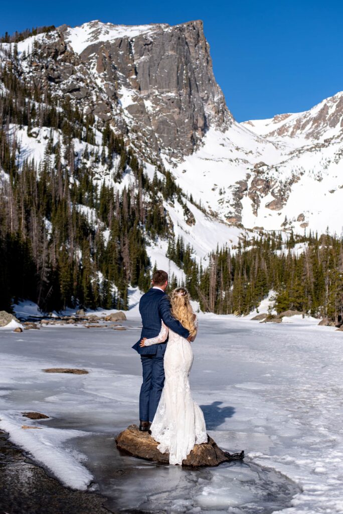 A bride and a groom are standing on a rock in the middle of a frozen lake looking away from us at a rock face covered in snow. The bride is wearing a long sleeved wedding dress and the groom is wearing a blue suit. 
