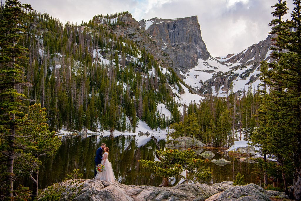 A bride and groom are standing on a rock with a lake and a mountain range behind them. There is snow on the ground and the couple is wearing wedding attire. 