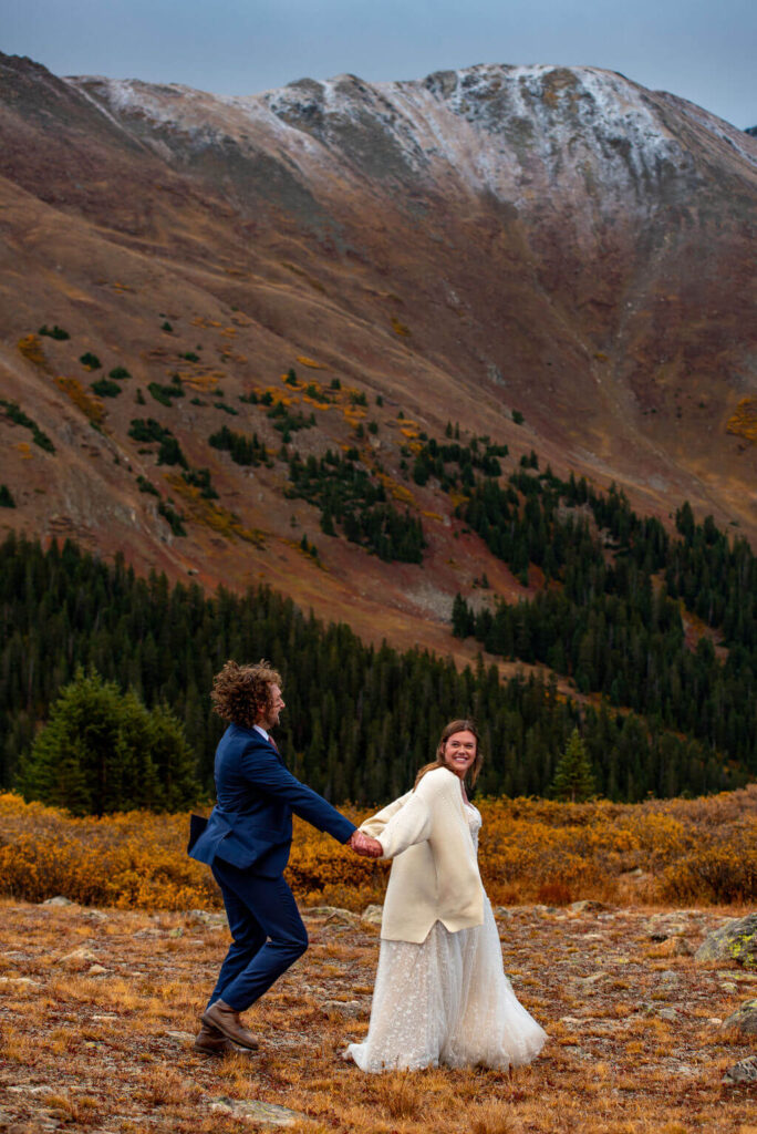 A bride in a white wedding dress and the groom in a blue wedding suit are running through a field with snowcapped mountains in the background. The couple is holding hands and the bride is looking back at the groom. 
