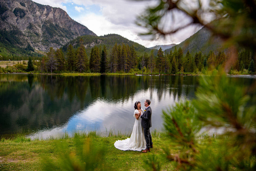 A man and a woman in their elopement outfits are dancing on the edge of a lake with trees and mountains in the background. In the foreground there are branches and leaves from a tree. 