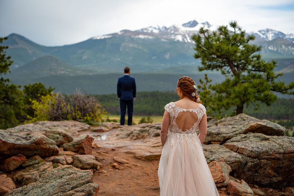  A bride is walking away from the camera towards the groom who is standing in the background. They couple is prepared for a "first look" before their elopement ceremony. There are mountains in the background. 