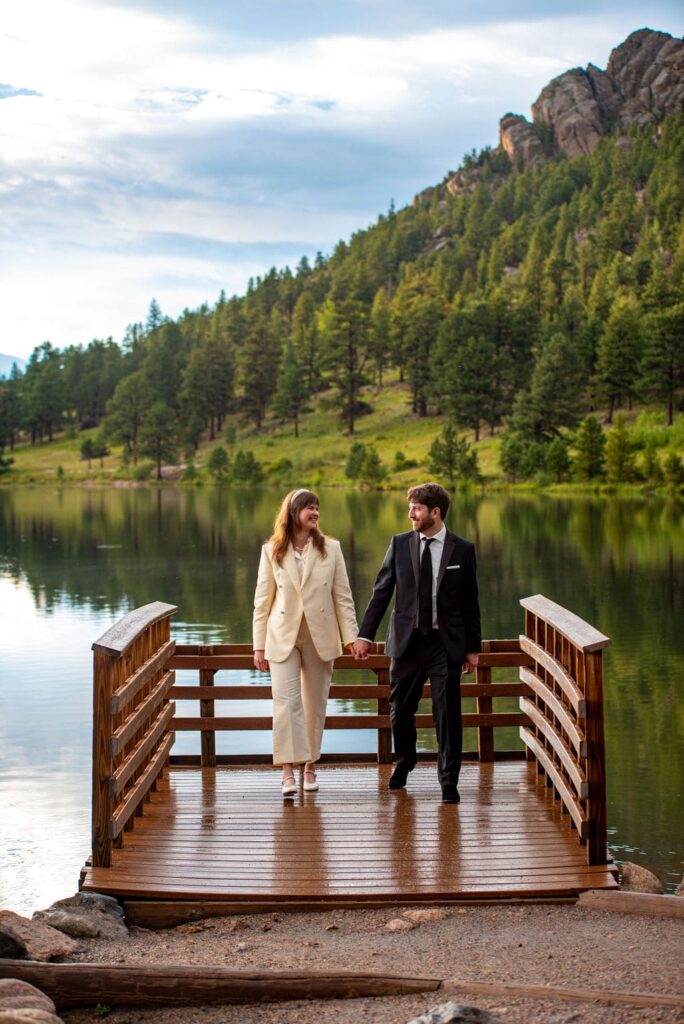 A woman in a white pants suit and a man in a suit and tie are holding hands and walking towards the camera. They are walking on a brown dock that sits on top of a lake with a green hill in the background. 