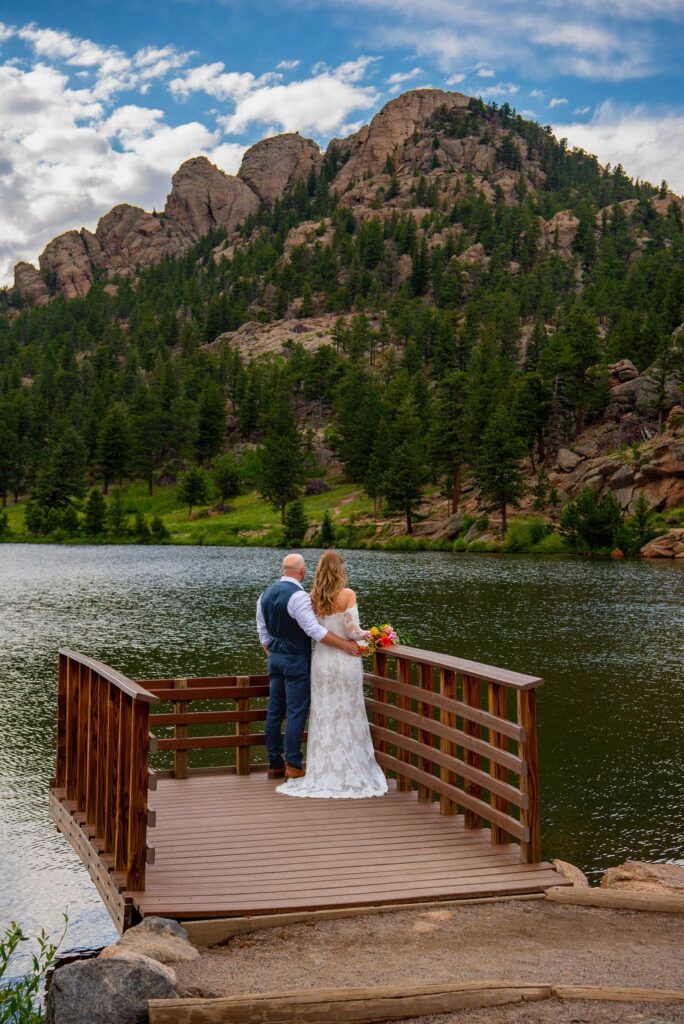 A bride and groom stand on a wooden dock overlooking a lake. The couple is facing mountains in the background. The man has his arm around the bride who is in her sleeved lace wedding dress.