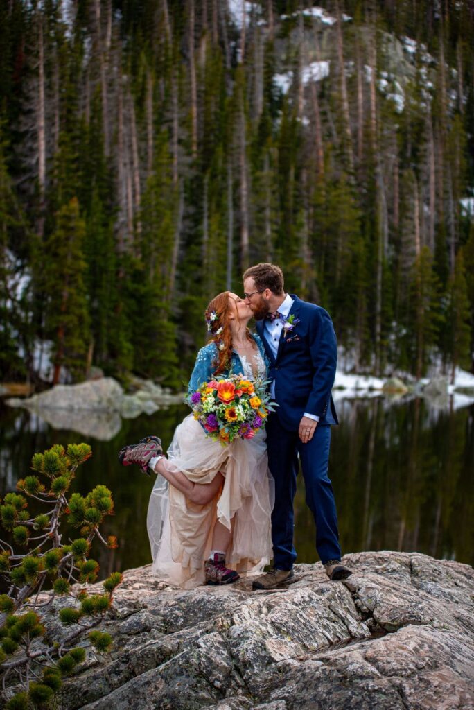  A bride in a wedding dress is wearing a jean jacket, holding a bright bridal bouquet and is kissing the groom. They are showing off their hiking boots while standing on a rock that is above a lake. 