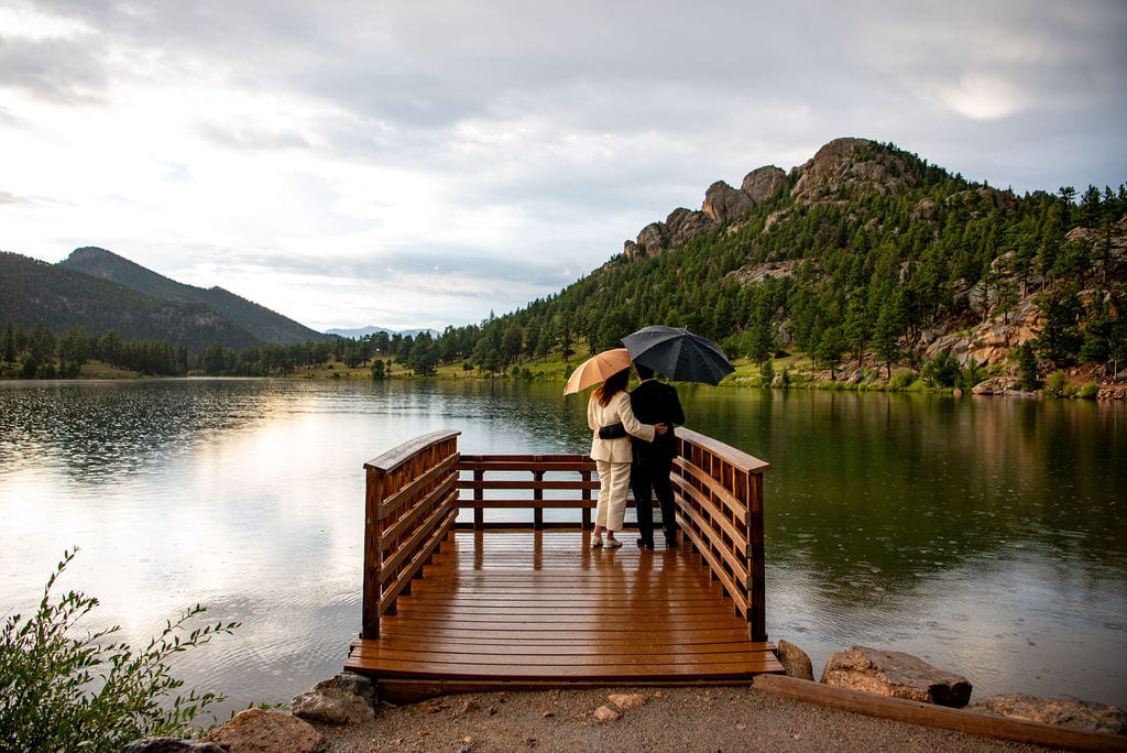 A man and a woman are standing on a brown dock with umbrellas. The dock looks out at a lake and mountains in the background. It is raining and you cn see ripples in the water.