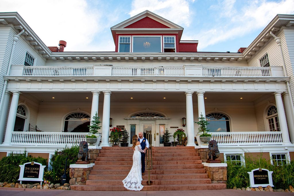A bride in her wedding dress is standing with her groom on a set of stairs in front of one of the buildings at the Stanley Hotel in Estes Park. The building is white with red accents. The couple just had a rocky mountain elopement. 