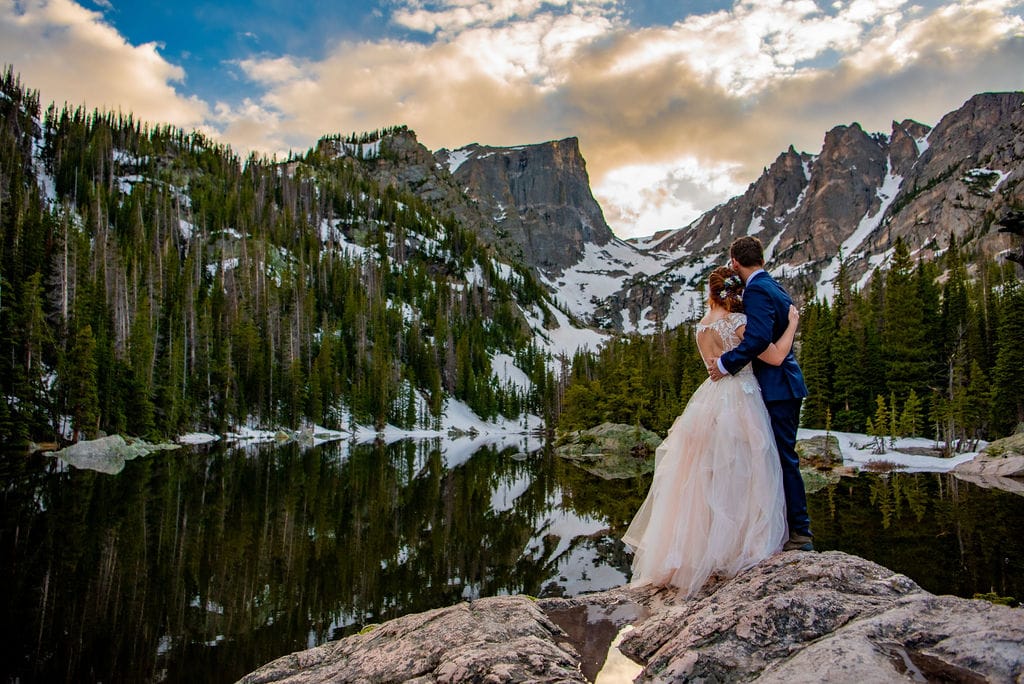 A bride and groom stand in their wedding attire with their arms around each other. They are facing away at the mountain range in the background. They are standing on a rock at Emerald Lake in Rocky Mountain National Park. The sun is setting behind the mountain, turning the sky yellow.