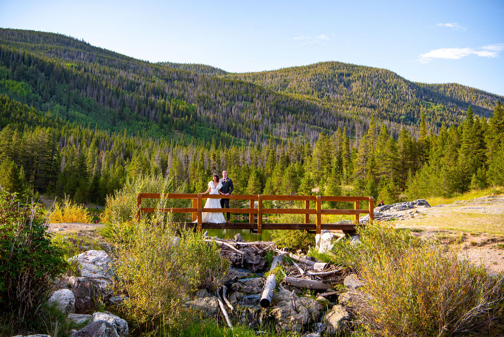 bride and groom standing on a wooden bridge surrounded by lush greenery and mountain views
