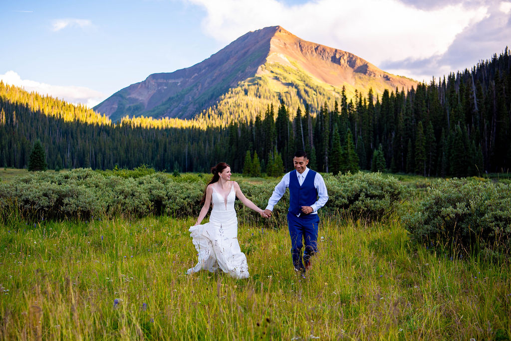 bride and groom running through a lush field holding hands with mountains in the backdrop for their Crested Butte elopement