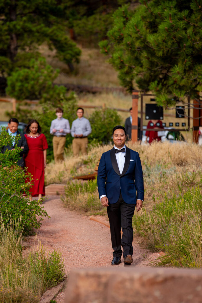 Groom in navy tuxedo walks down a nature trail lined with family at a beautiful Boulder Halfway House wedding.