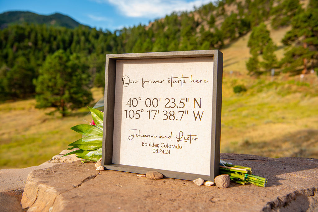 Framed sign with elopement coordinates and wedding date placed on a rock with scenic Boulder, Colorado mountains in the background. The sign reads “Our forever starts here,” perfect detail captured by a Boulder Colorado elopement photographer.