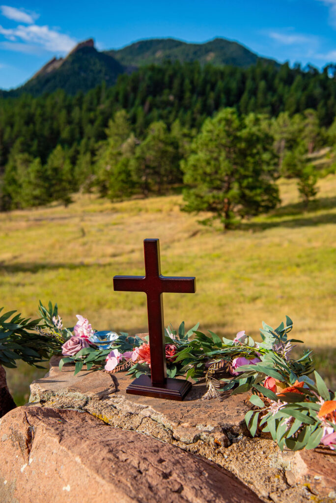 Wooden cross and floral arrangement on the stone altar at a Boulder Halfway House wedding with views of the surrounding mountains.
